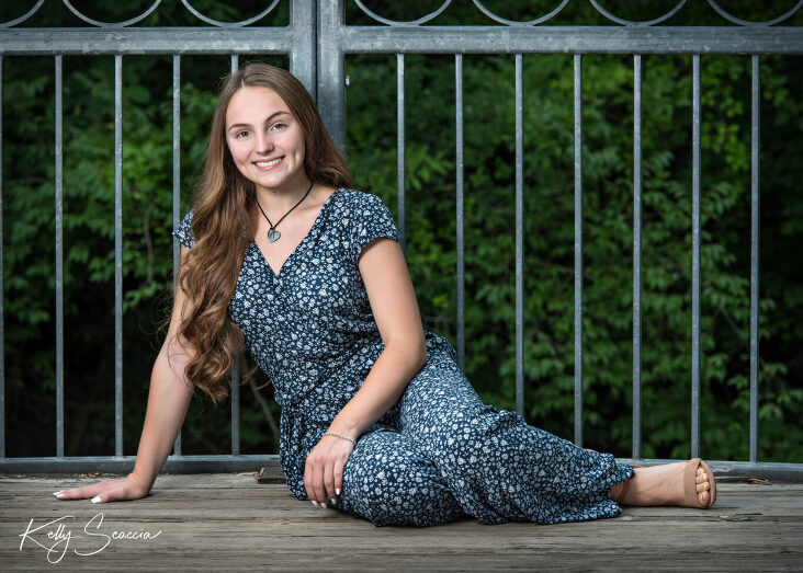 Girl senior portrait outdoor smiling looking at you in a blue and white short sleeve pantsuit iron rails and greenery behind her sitting on floor