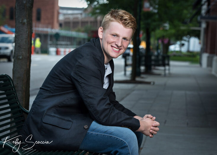 Senior guy outdoor city portrait wearing jeans, gray tshirt, black sport coat smiling looking at you sitting on a bench