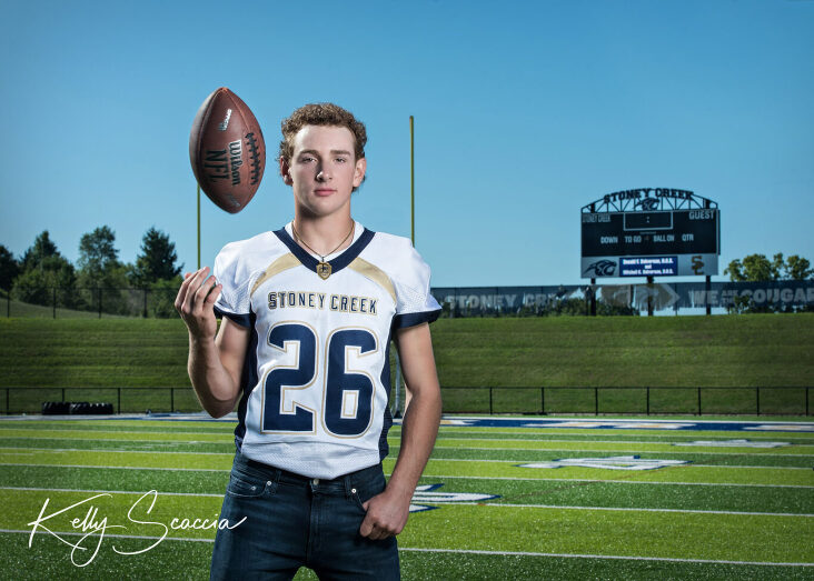 Senior guy outdoor football portrait wearing football jersey and jeans looking at you on the football field tossing the football in the air