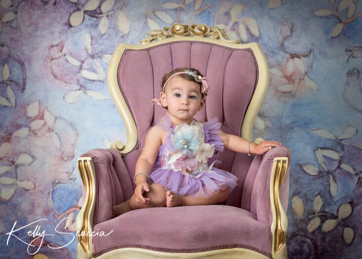 Studio one year little girl portrait in a purple tutu outfit looking at you with a serious expression sitting on a purple chair 
