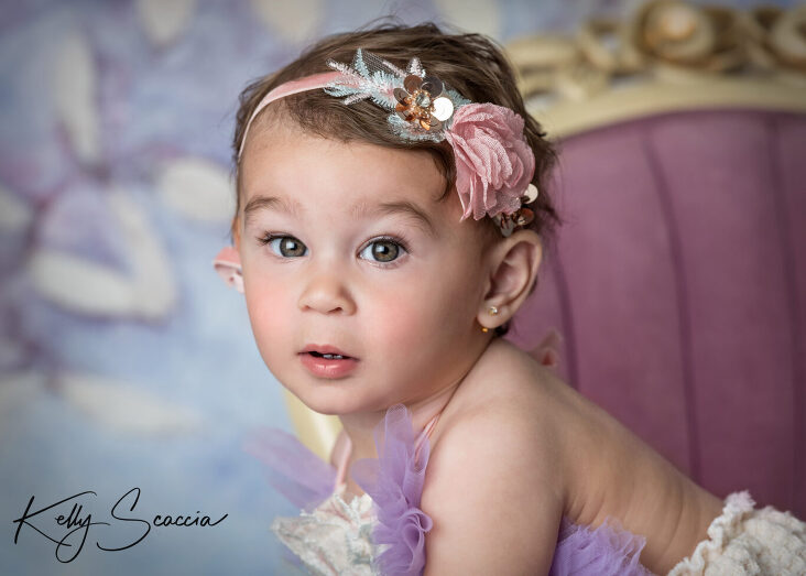 Studio one year little girl portrait in a purple tutu outfit looking at you with a serious expression sitting on a purple chair headshot