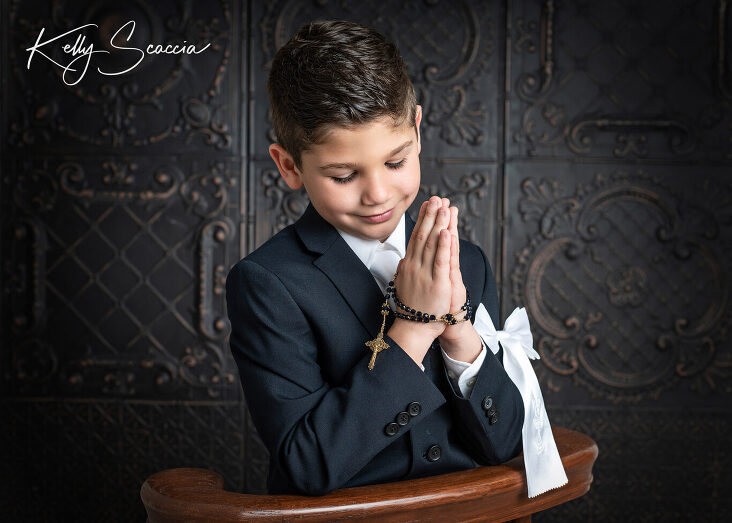 Studio communion boy portrait wearing a dark, navy suit, white bow on arm, holding his rosary, smiling, looking at his hands