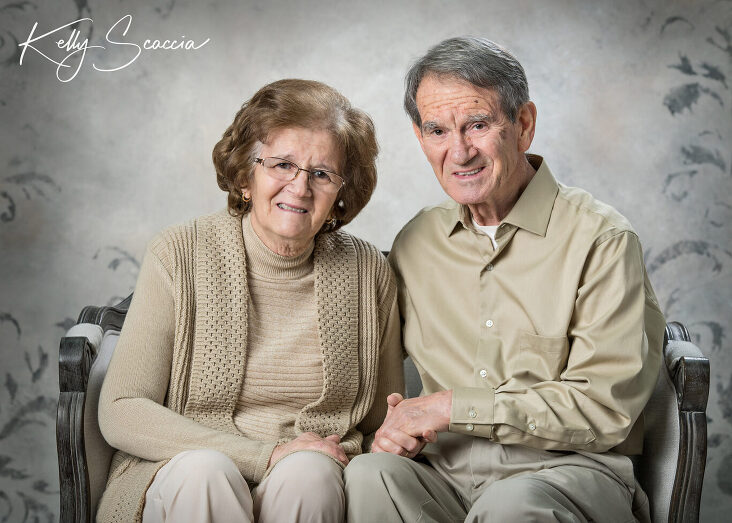 Studio portrait of grandma and grandpa, wearing brown tones, sitting on a couch, smiling looking at you