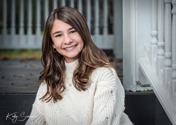 Outdoor portrait of teenage girl, long dark hair, brown eyes, looking at you, smiling, wearing cream sweater