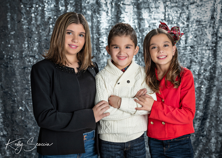 Studio portrait of two tween girls in black and red and jeans with little brother in cream sweater and jeans, all dark hair, dark eyes, looking at you, smiling