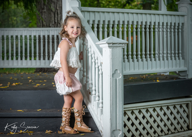 Outdoor little girl portrait in white shirt, pink bloomers, tall gladiator boots, curly light hair, light eyes, in a gazebo, looking at you, smiling