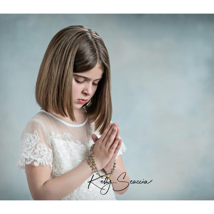 Studio communion girl in white dress portrait, straight brown hair standing with rosary in her hand looking down 