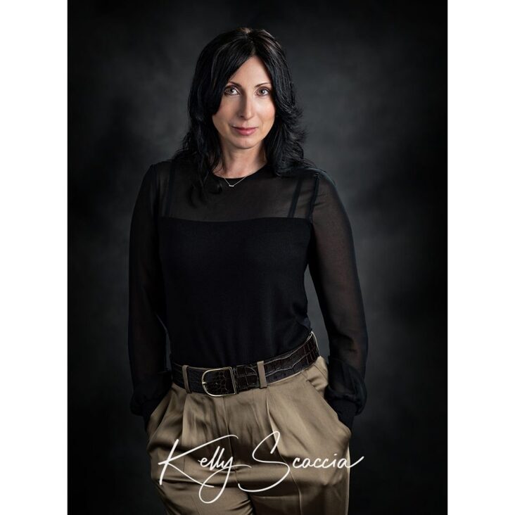 Studio portrait headshot woman with black hair, dark eyes, looking at you, smiling, wearing black shirt and tan pants