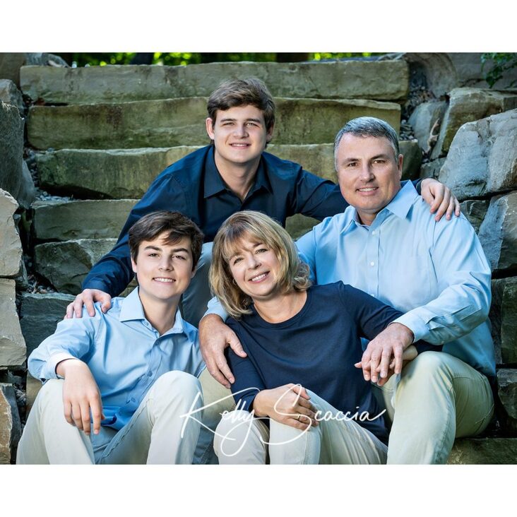 Outdoor family portrait of mom, dad and two grown sons wearing blue shirts, tan pants, looking at you, smiling