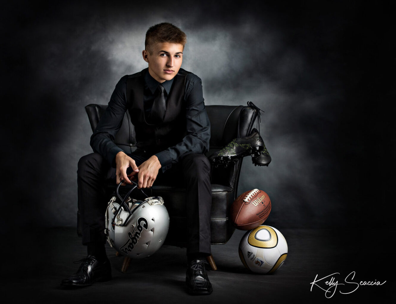 High School senior guy in studio wearing black suit sitting in chair with football helmet, cleats, football and soccer ball