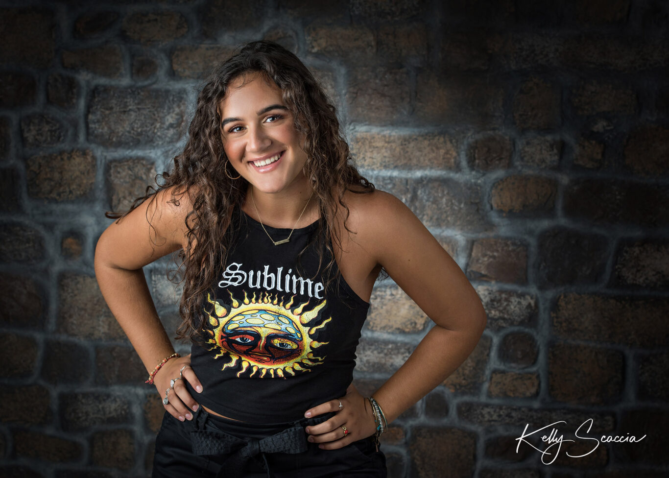 High School senior girl studio portrait in front of a brick wall wearing a Sublime sleeveless shirt smiling with hands on hips