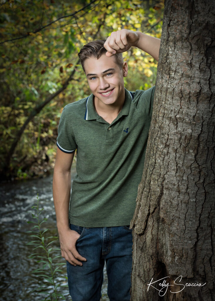 High School senior guy outdoor in the park with hand against a tree leaning forward smiling