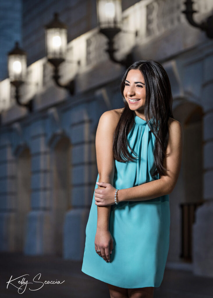 High School senior girl in front of white marble museum smiling wearing a teal dress holding her arm looking off to the side