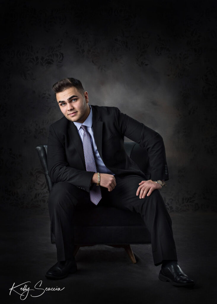 High School senior guy in studio sitting on a chair leaning on thigh serious expression