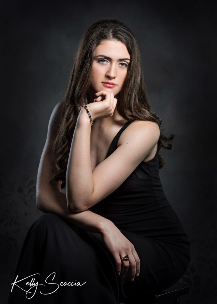 Studio portrait on dark background of tall girl with long, dark hair wearing a fitted, long black dress sitting on a stool looking straight at you with a serious expression while crossing her arms in lap and putting one hand under her chin