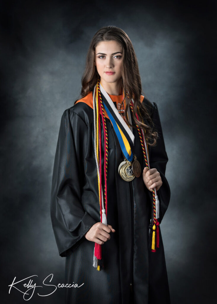 Studio portrait on a dark background of a tall girl with long, dark hair wearing a black graduation gown with layers of medals and awards looking straight at you with a serious expression