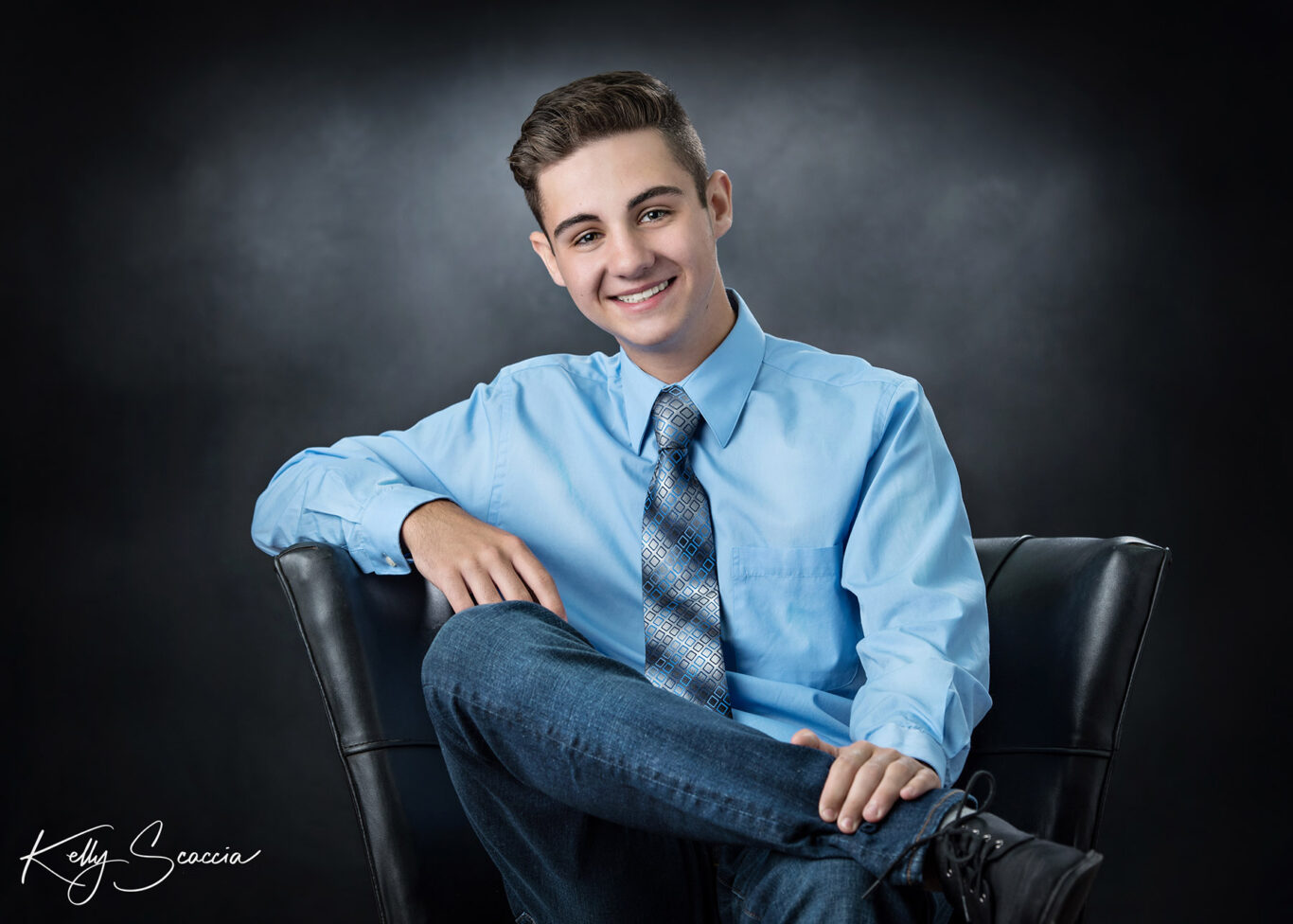 High School senior guy studio portrait sitting on black chair leaning back legs crossed smiling